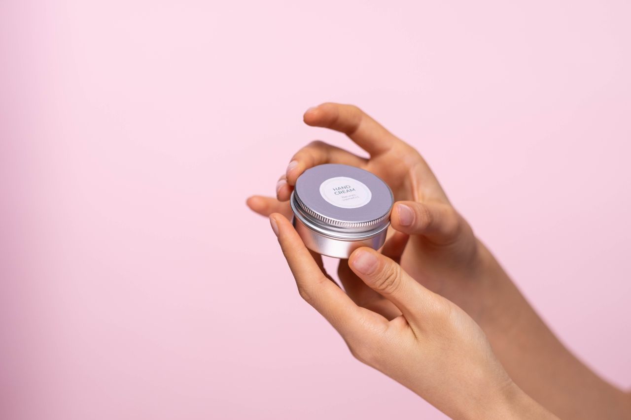 services-04 Close-up of a person holding a moisturizing hand cream jar against a pastel pink background.